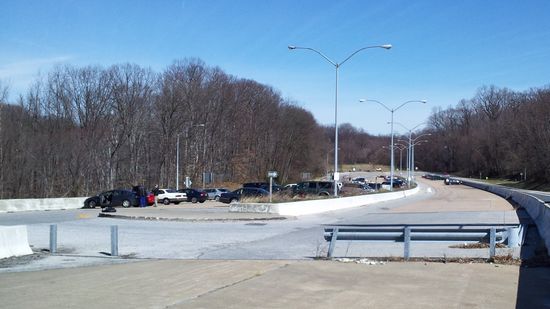 Overview of the I-70 park and ride from the western end of the facility. The intended westbound lanes (where most of the cars are parked) are to the left of the barrier in the center of the photo, while the intended eastbound lanes are to the right of said barrier.