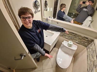 Elyse shows off the one-piece toilet in the bathroom, while I am visible in the mirror taking the photo.