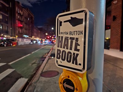 "HATE BOOK" sticker on a sign for a pedestrian signal call button at the intersection of Carmine and Varick Streets.