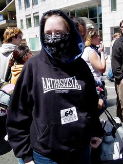 A masked woman wears an anti-fascist sweatshirt, while also wearing a "60 Years of Destruction" sticker. Sis and I also wore these stickers, which were being given out at the rally site.