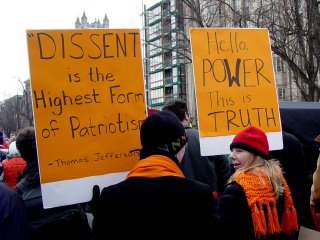 As one sign reminds us what patriotism really is, the other speaks truth to power.