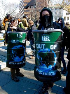 These shields, which read "U.S. OUT OF EVERYWHERE", were made of 90-degree sections of a plastic barrel. They were painted with the green-and-black flag of green anarchism as a background, overlaid with a picture of the Earth showing the eastern hemisphere and the lettering. Then they had padding added to the top, and a neck strap. These shields often held the front spot in the march.