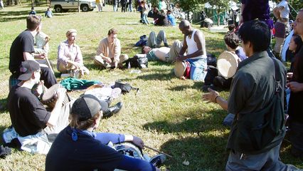 A group sits on the grass north of the reflecting pool, playing real drums, as well as tambourines.