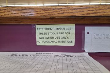 Sign on a seating area advising employees that the stools are intended for customer use only.  Clearly, no one pays attention to this sign, because based on what I found there, someone had been using it to work on employee schedules in that seat.