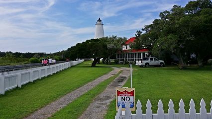 Ocracoke Lighthouse in the distance, with the lighthouse keeper's house in front. The keeper's house is now a private residence. The raised walkway is visible at far left, beyond the white fence.