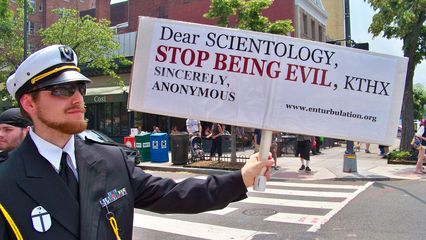 John McNonymous, dressed in a manner resembling a naval dress uniform, holds a sign saying, "Dear Scientology, stop being evil, KTHX. Sincerely, ANONYMOUS."