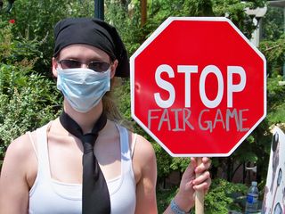 A woman wearing a necktie and a surgical mask holds a sign saying "STOP FAIR GAME".