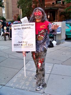 With a new twist on the lighted-eyes theme, this Anon dressed as a zombie, with a sign saying, "I joined Scientology for the BRAINS... but they DON'T HAVE ANY." The shirt says "Billion year contract" on the front, and "Die-enetics: Get it... read it... brainwash it!" on the back.