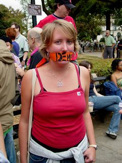A woman is symbolically gagged (with "democracy" written on the orange cloth) while also wearing a necklace with a less-than-flattering message about George W. Bush on it.