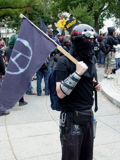 Black bloc demonstrators' statements took various forms. The gentleman on the left carries a flag, flown upside down (an international distress signal), with "smash the state" written on it. Another gentleman is dressed for battle, wearing a helmet, goggles, padding on both arms, and carrying an anarchist flag.