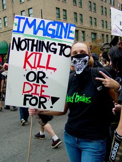 A woman wearing a bandanna with part of a skull printed on it carries a sign saying, "Imagine nothing to kill or die for!"