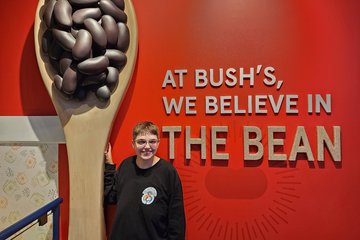 Elyse poses with a giant spoon holding some giant beans, along with lettering that says, "At Bush's, we believe in THE BEAN".  The letters in the last two words, meanwhile, are filled with beans.