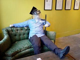 The mannequin of the police officer depicts police in an unfavorable light, showing a somewhat-overweight officer sitting on a couch watching TV, with one foot on the coffee table, nightstick tucked under one arm, shirt untucked, and wearing a riot shield and graduation cap.
