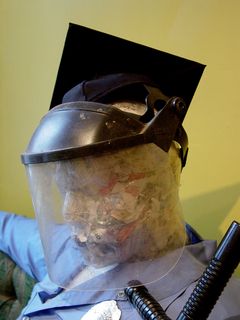 The mannequin of the police officer depicts police in an unfavorable light, showing a somewhat-overweight officer sitting on a couch watching TV, with one foot on the coffee table, nightstick tucked under one arm, shirt untucked, and wearing a riot shield and graduation cap.