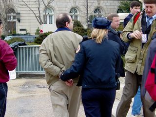 When I first arrived, two people were engaging in civil disobedience. It was stated by law enforcement officers on site to stay on the sidewalk, and not to go onto the steps. When these individuals stepped off the sidewalk and onto the Supreme Court's patio, law enforcement took notice. Pete Perry, wearing the brown jacket, was detained briefly and released (the officer is merely holding his hands behind his back), while Midge Potts, wearing a green shirt and an Abu Ghraib hood, was handcuffed and arrested.
