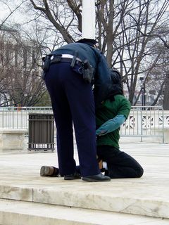 When I first arrived, two people were engaging in civil disobedience. It was stated by law enforcement officers on site to stay on the sidewalk, and not to go onto the steps. When these individuals stepped off the sidewalk and onto the Supreme Court's patio, law enforcement took notice. Pete Perry, wearing the brown jacket, was detained briefly and released (the officer is merely holding his hands behind his back), while Midge Potts, wearing a green shirt and an Abu Ghraib hood, was handcuffed and arrested.