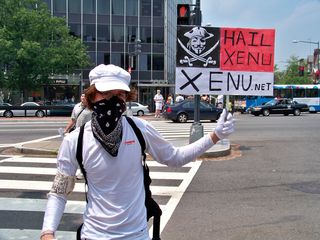 A masked individual holds a sign advertising the Web site Xenu.net.