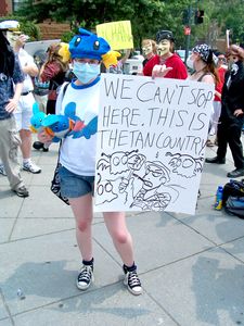 A woman wearing a surgical mask and decked out in mudkips holds up a "Thetan country" sign.