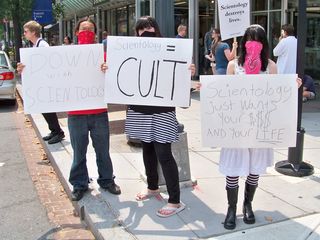 Three masked individuals hold signs critical of the Church of Scientology.