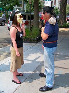 A woman in a Guy Fawkes mask carries on a conversation with Arnie Lerma on the sidewalk in front of the Founding Org.