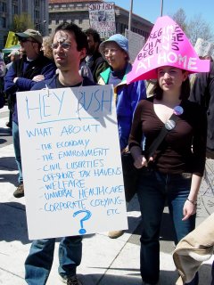 A number of more pressing issues are at hand, as evidenced by the sign held by the gentleman at left. Additionally, he has a peace sign painted on his forehead. The woman next to him has an oversized hat, stating that regime change begins at home, implying that Bush should resign.