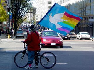 Ahead of the group, a woman on a bicycle holds a flag featuring a dove with a rainbow streaming behind it.