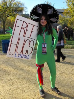 A woman wearing a green zentai (with the hood down) and a giant hat holds up a sign offering "Free Hugs for Liberals".