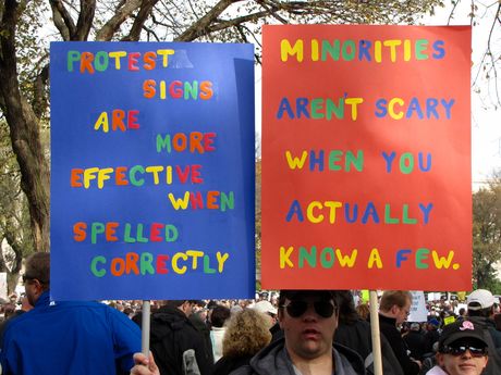 A man holds two signs - one reminding everyone why it's important to spell the messages on your protest signs correctly, and the other about minorities.