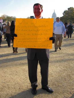 A man spoofs then-House Minority Leader John Boehner with an orange-painted face, and a sign encouraging viewers to guess who he is supposed to be.