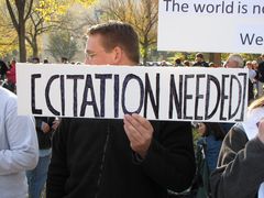 A man holds a sign containing the {{citation needed}} template from Wikipedia.