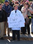 A man wearing an Aperture Science (from Portal) lab coat holds a sign reading, "Fight Science with Wood!"