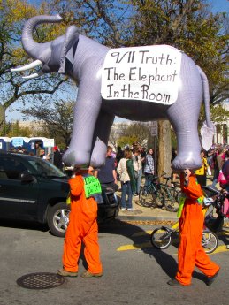Two people in orange jumpsuits carry a large inflatable elephant with banners discussing the 9/11 Truth movement.
