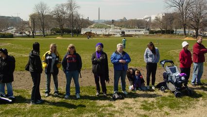 Meanwhile, just beyond a retaining wall, a small but determined group of people holds a silent vigil in protest of the occupation of Iraq on the year anniversary of the US invasion.