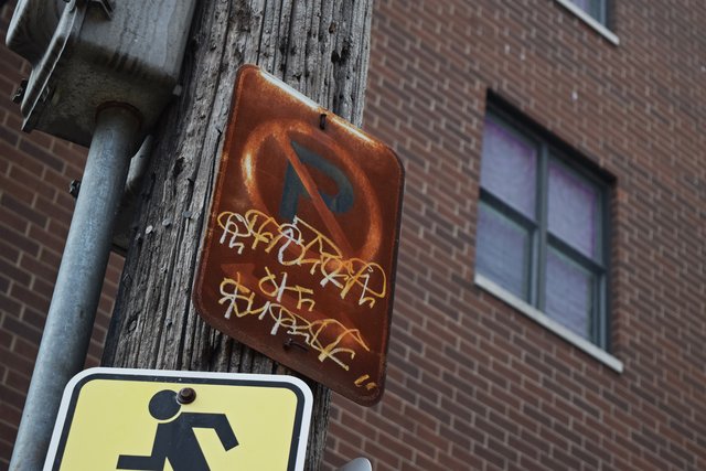 Graffiti covers the lower half of a rusted no-parking sign attached to a utility pole.