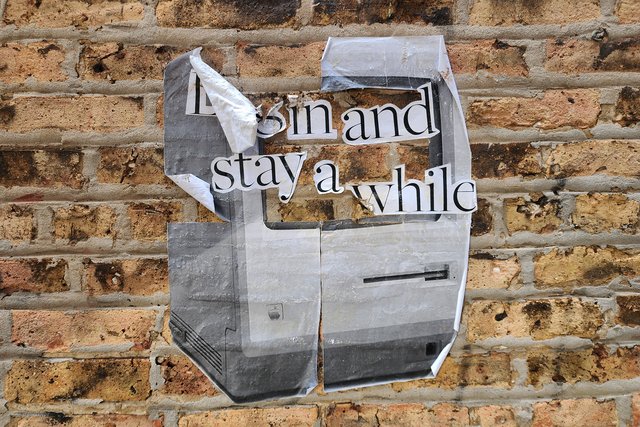 Weathered paste-up on a brick wall, featuring a classic Macintosh computer coupled with the line "Log in and stay a while."